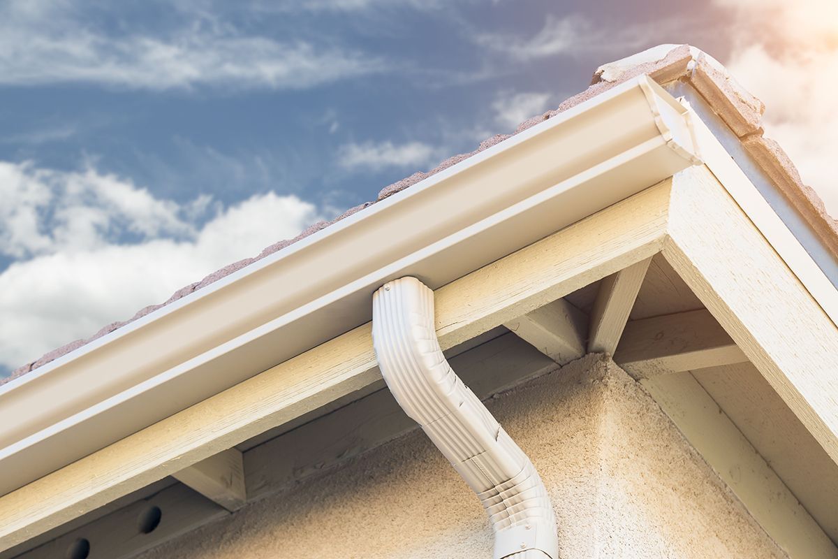 White rain gutter and downspout on a house roof against a cloudy sky