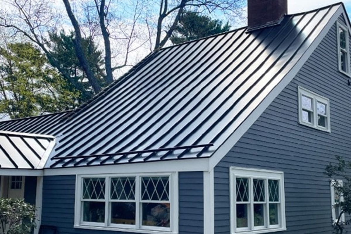 Gray house with a metal roof, white-trimmed windows, and a brick chimney under trees