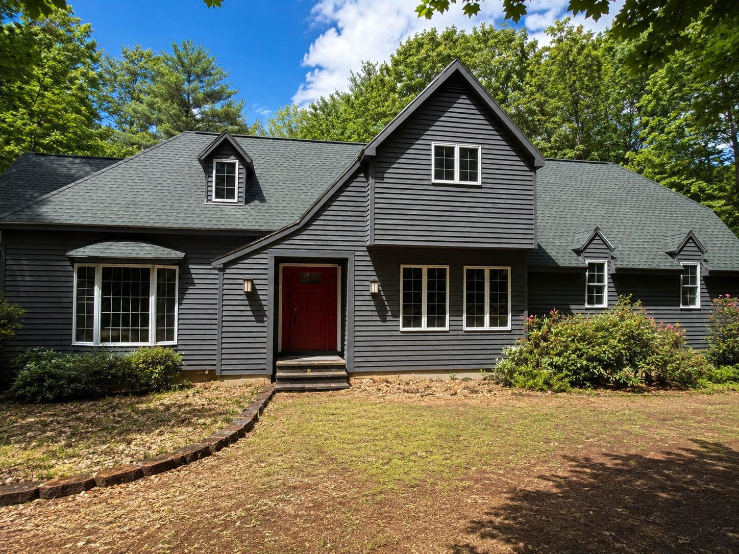 Gray house with red front door and dormer windows, set among trees and a leaf-covered yard.