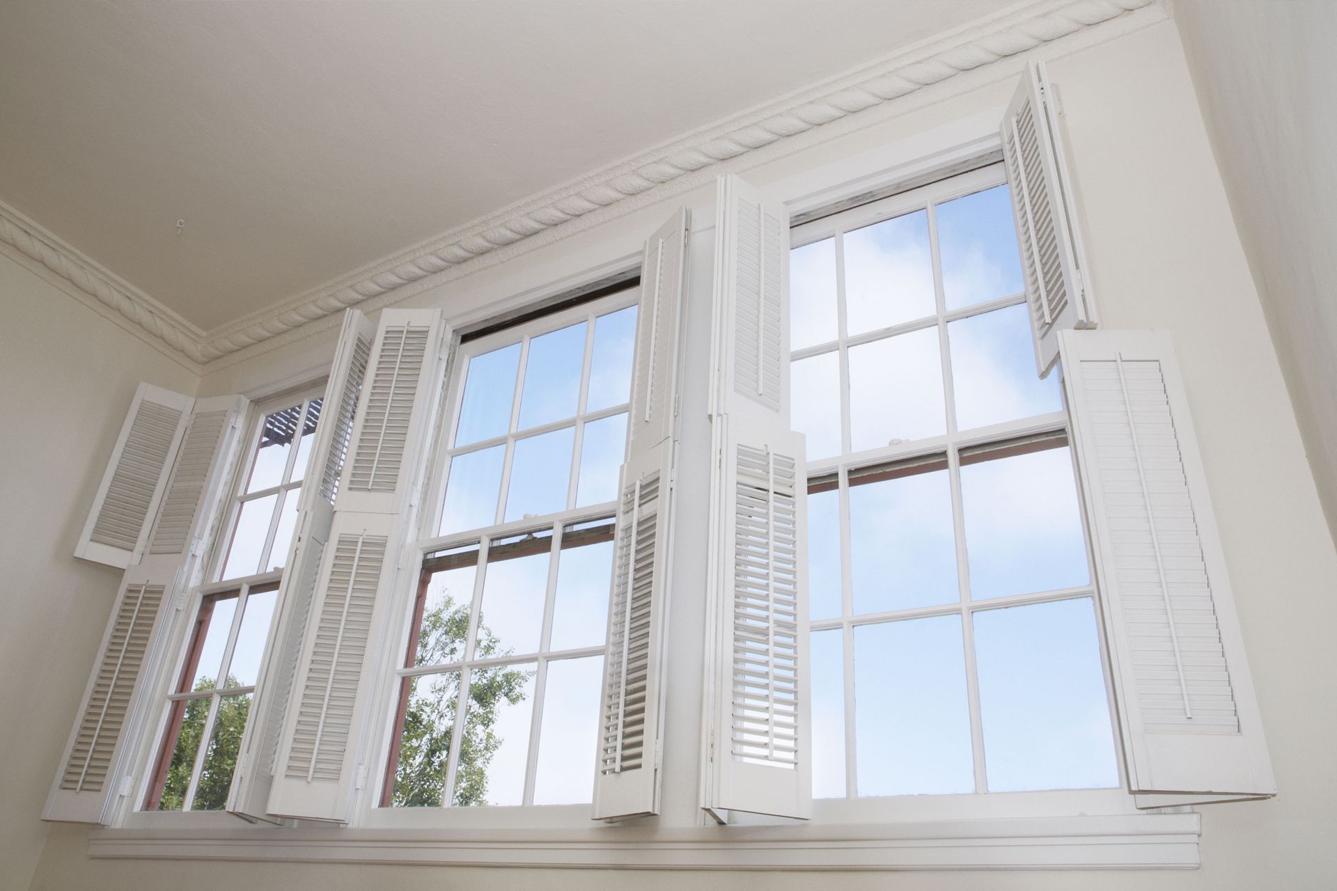 White windows with open shutters in a bright room, showing blue sky and trees outside.