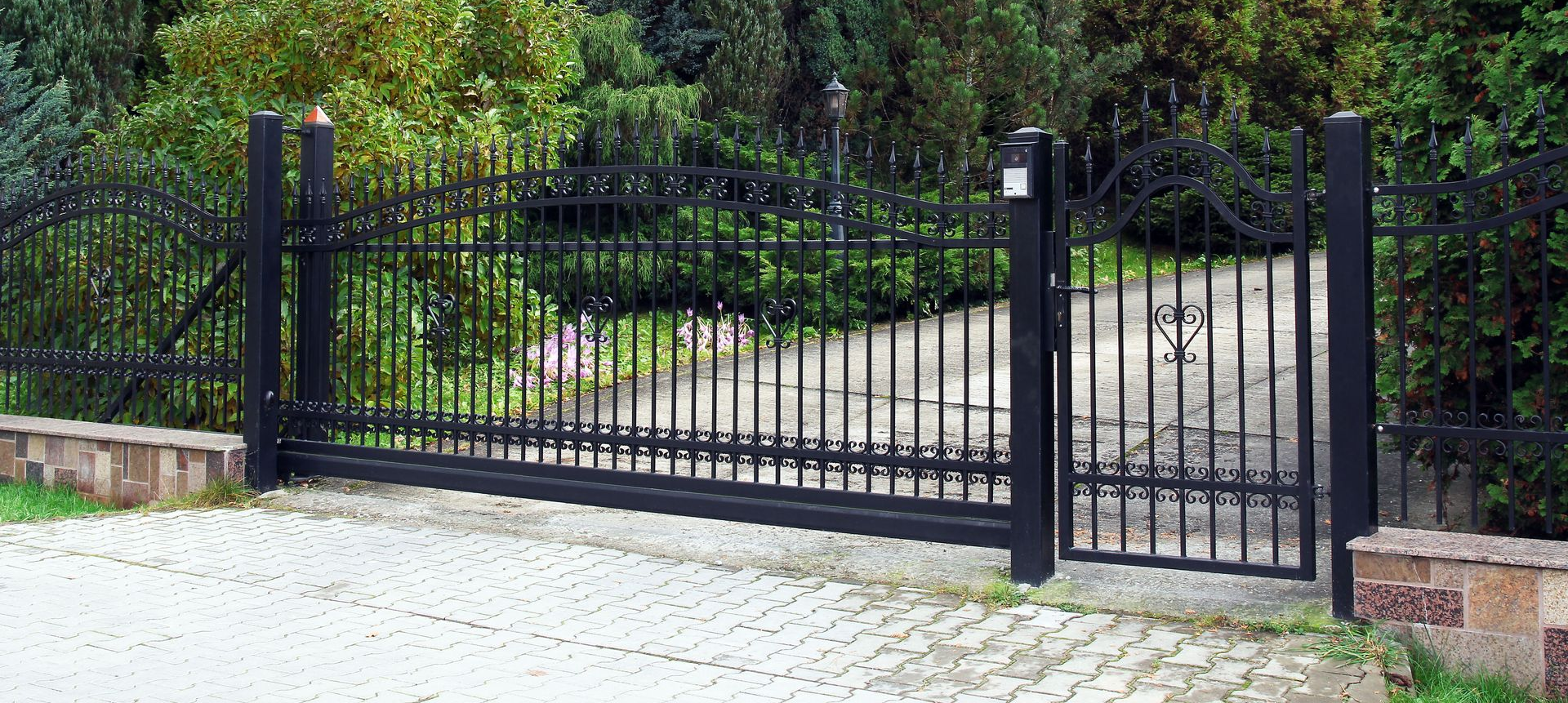 Black wrought-iron driveway gate between stone pillars, with greenery behind it.