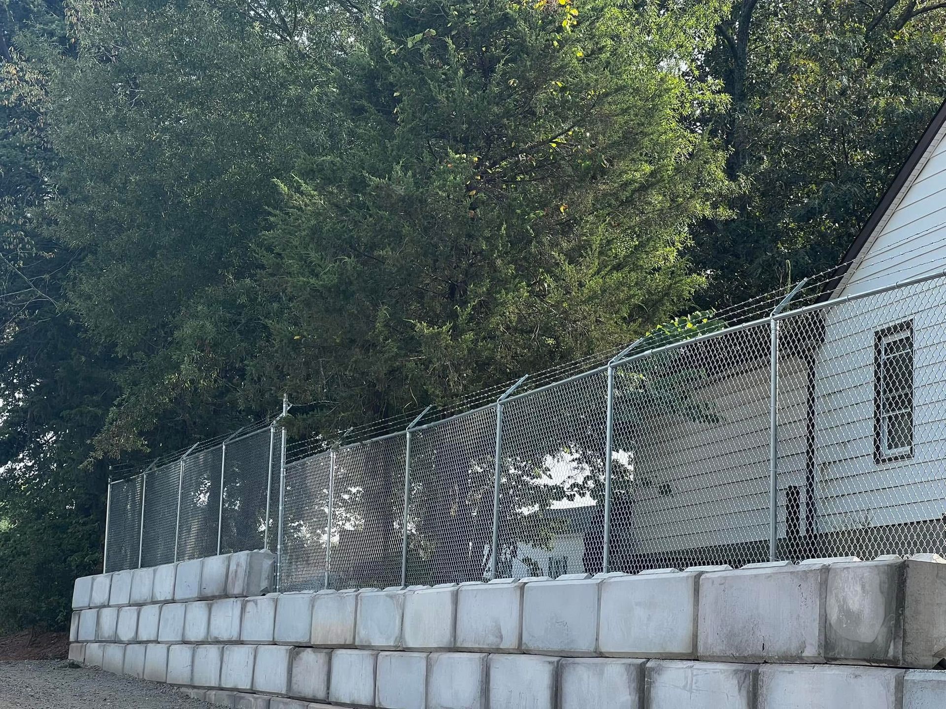 Chain-link fence and white retaining wall along a tree-lined hillside beside a small house.