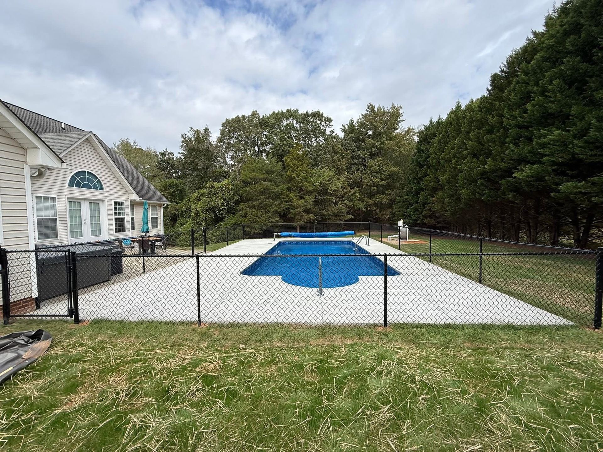 Fenced backyard with a covered swimming pool beside a house and trees beyond.