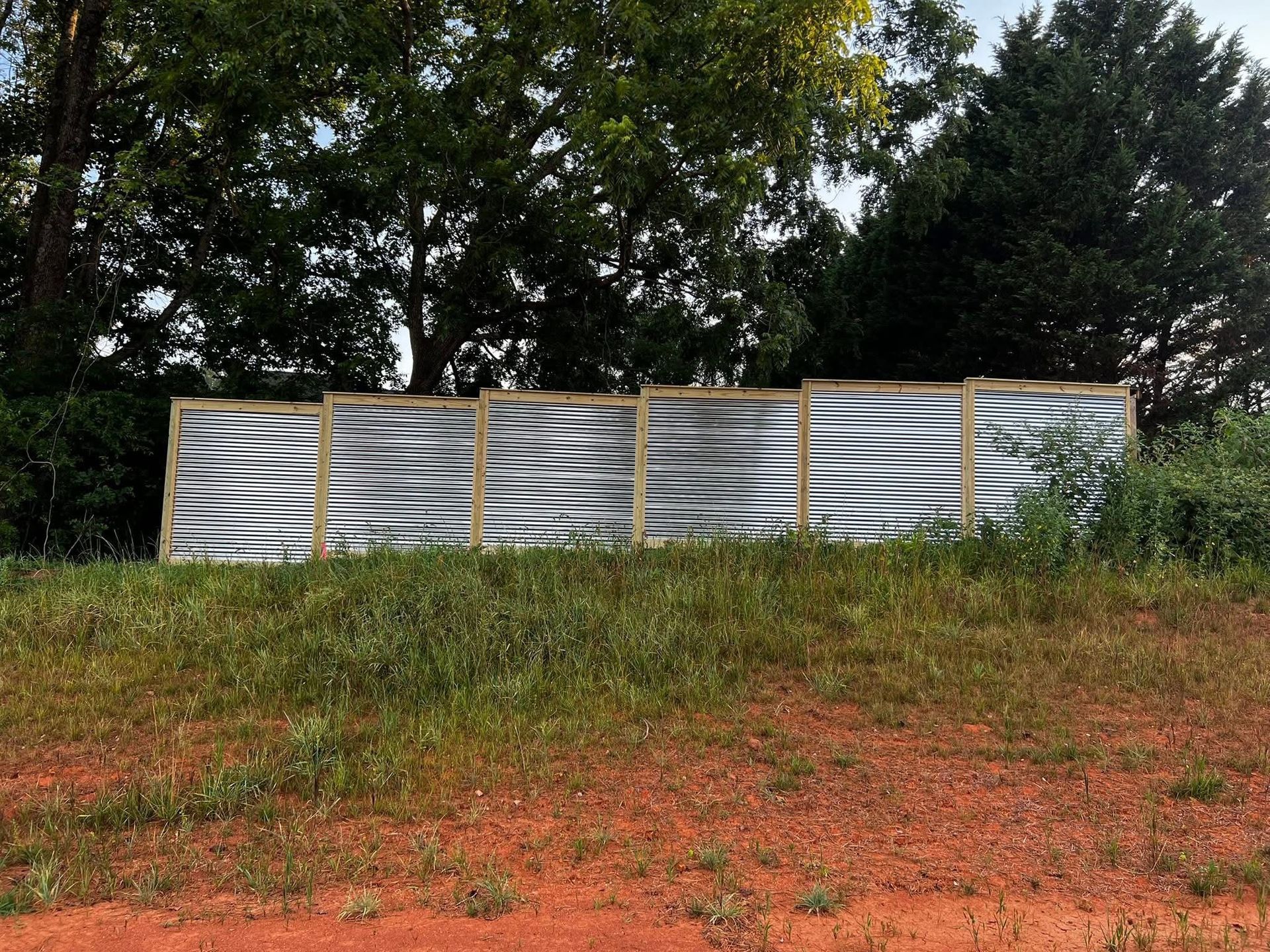 Corrugated metal fence panels along a grassy roadside, with trees behind and red dirt in front.