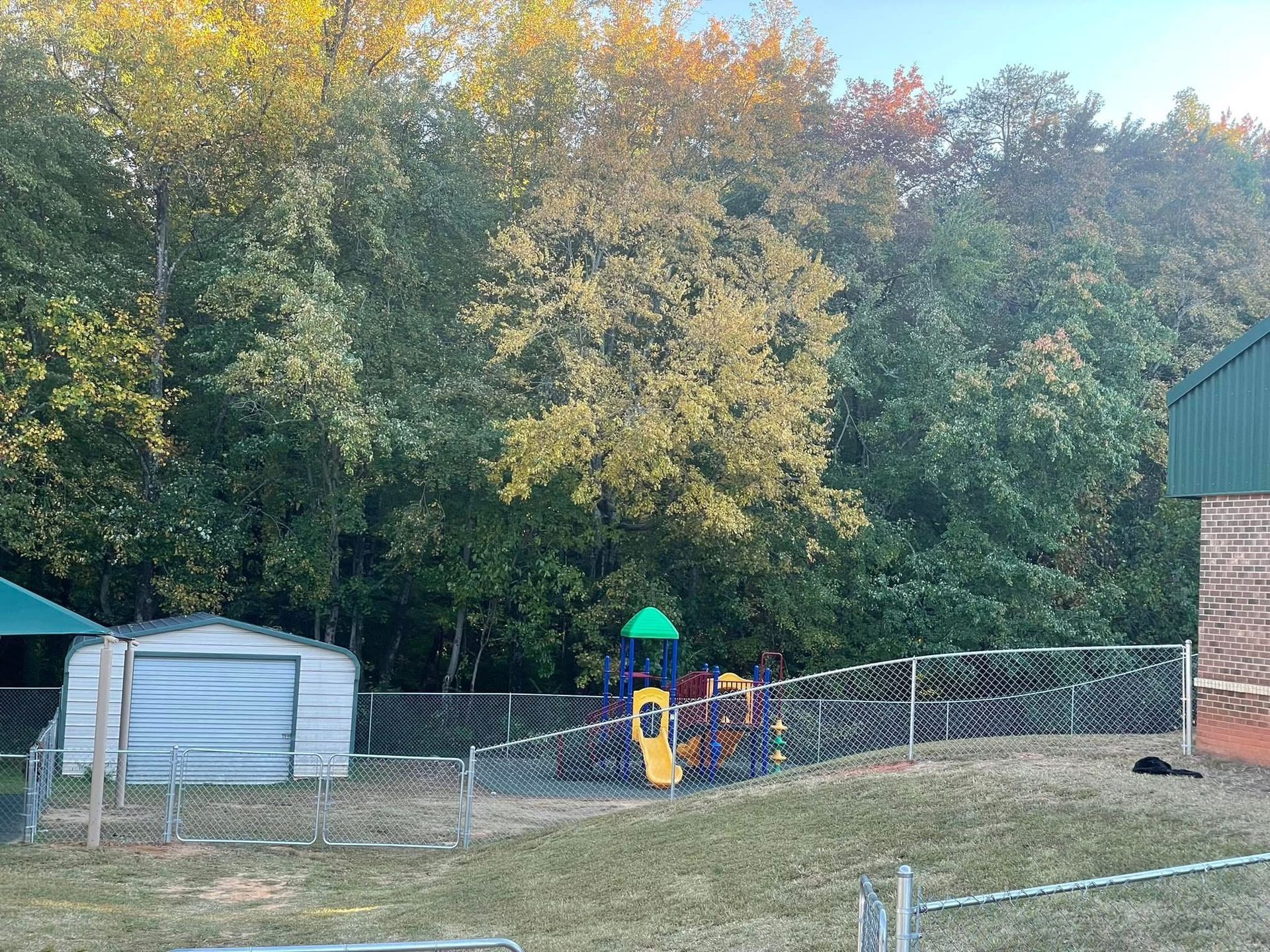 Small fenced playground with a play structure and shed, backed by dense autumn trees.