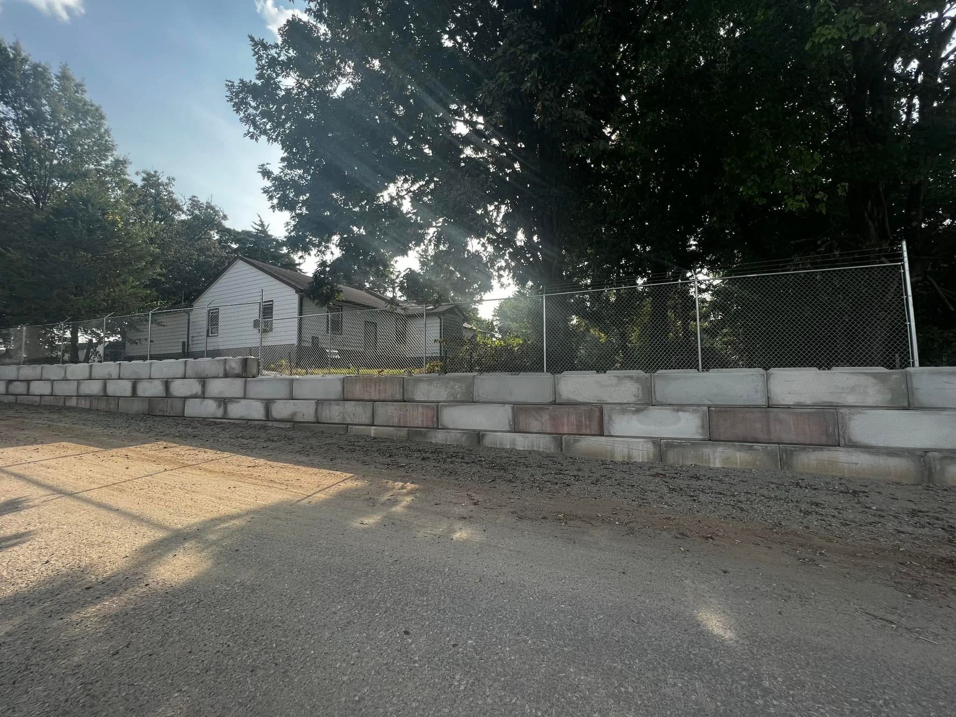 Small white house behind a low stone wall and chain-link fence, shaded by large trees on a roadside.