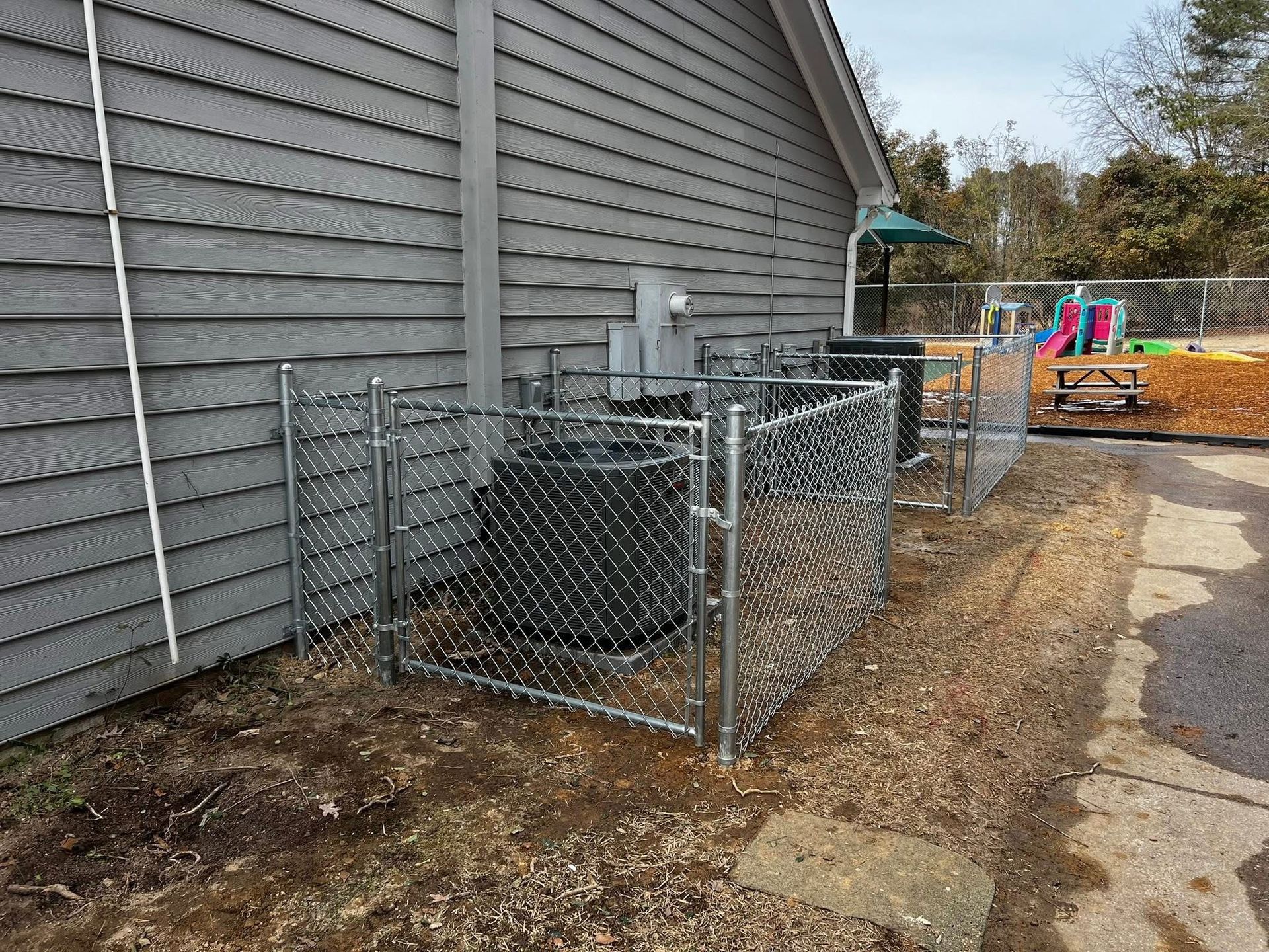 Chain-link kennel enclosures beside a gray house, with equipment and a dirt path in the yard.