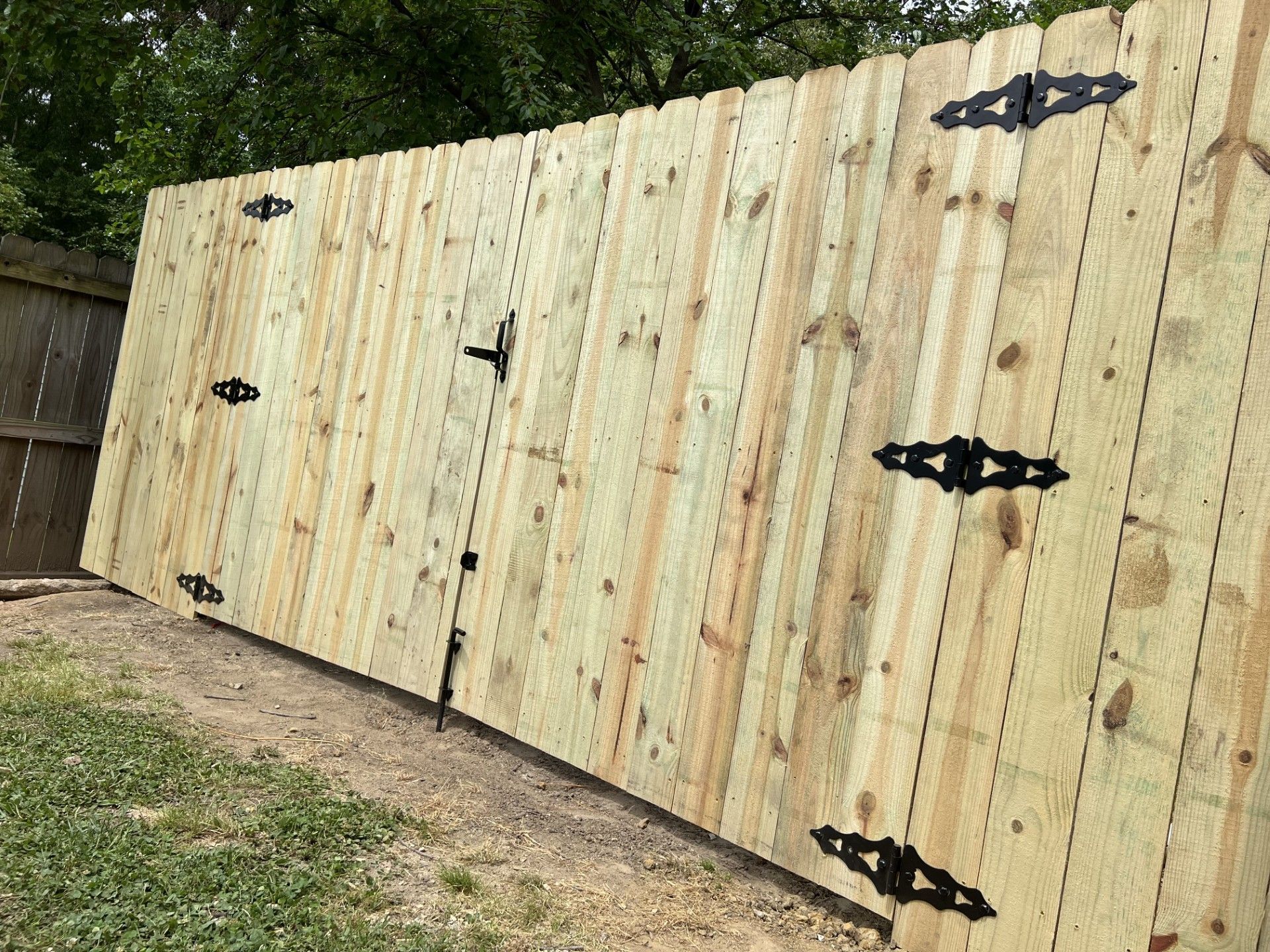 Tall beige wooden fence with black decorative hinges in a backyard, viewed at an angle.