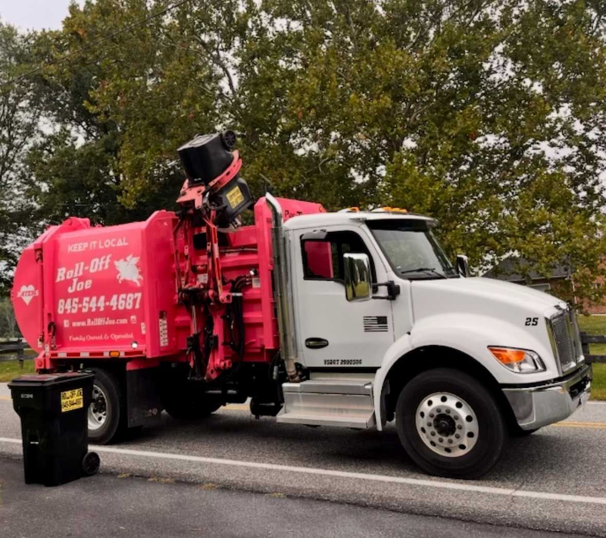 Pink and white garbage truck lifting a black trash can on a road.