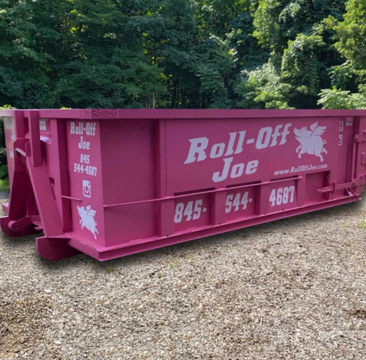 A bright pink dumpster sits on a gravel surface in front of green trees.