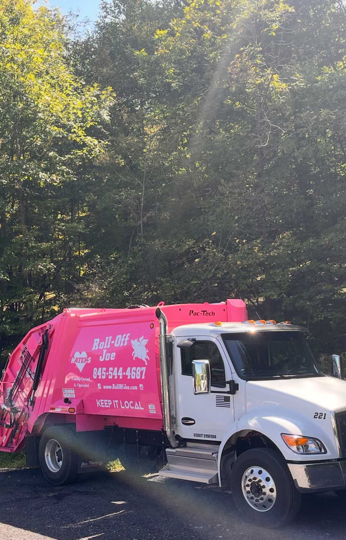Pink and white trash truck in front of trees. 
