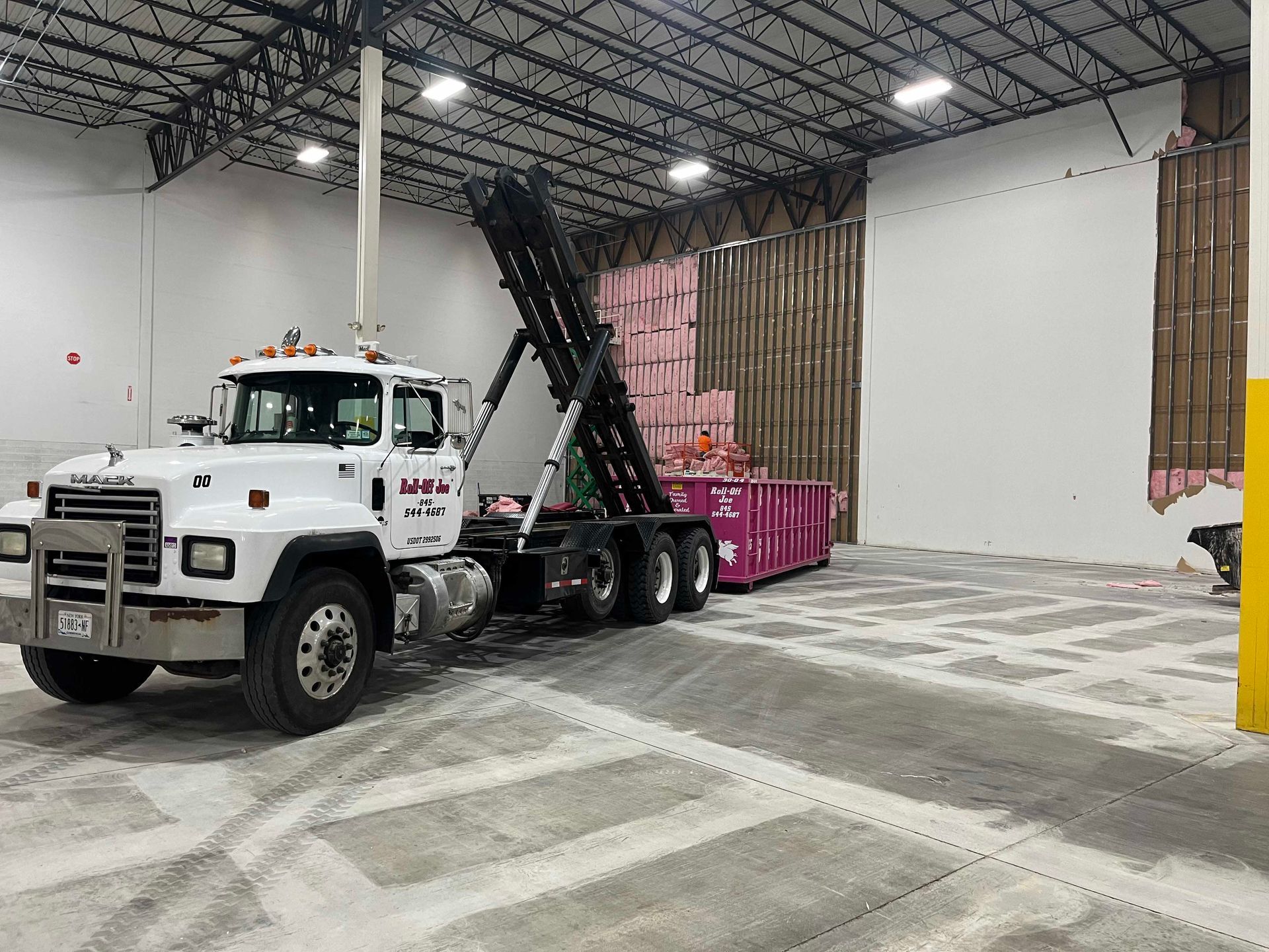 A white Mack truck with a pink dumpster inside a warehouse. The dumpster is loaded with pink insulation.
