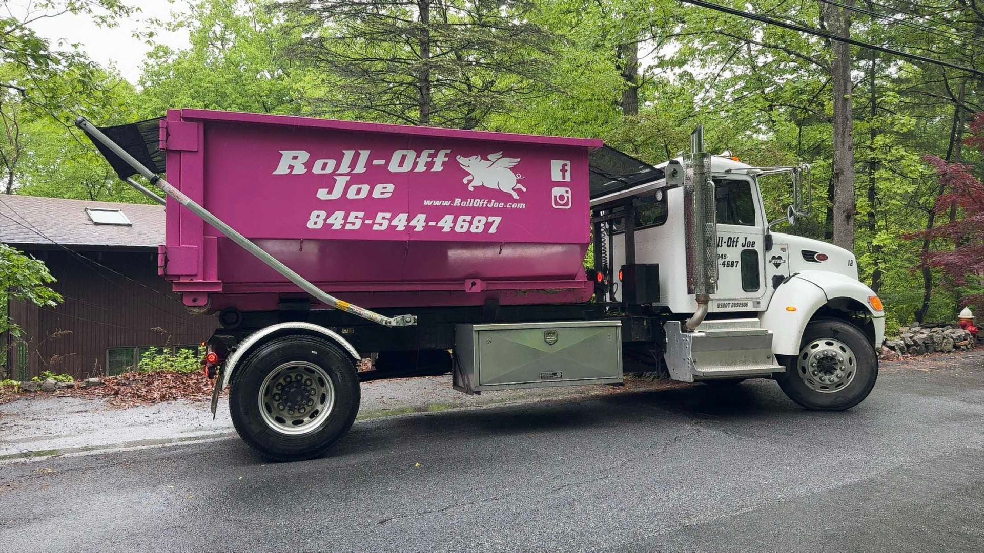 Pink Roll-Off Joe truck with container, parked on a road, trees in background, 845-544-4897.