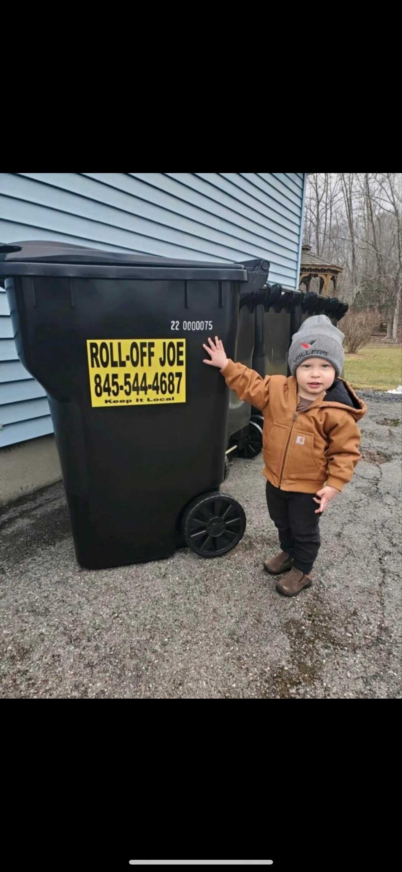 A young child wearing a beanie and jacket points at a trash dumpster labeled 