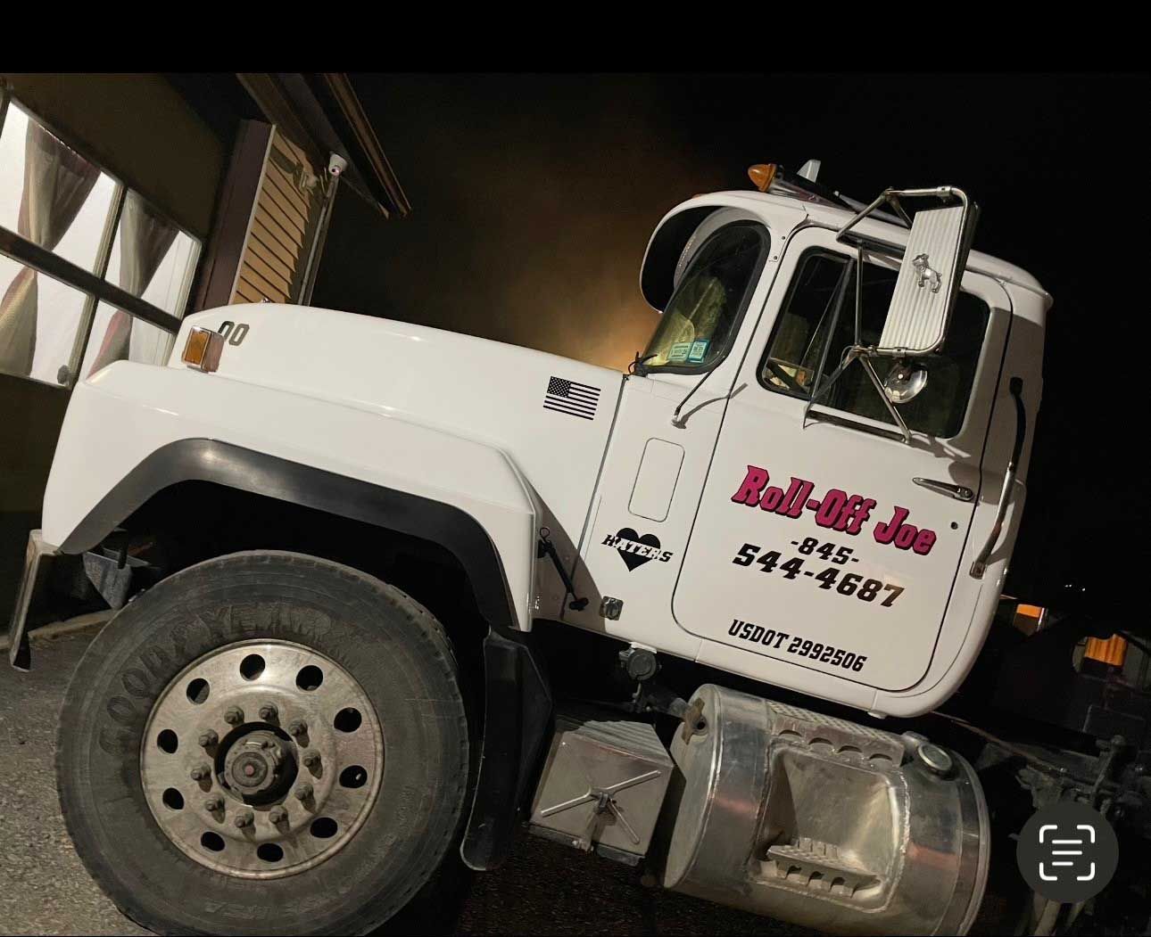 White Roll-Off Joe truck parked at night, with company logo and phone number on the side.