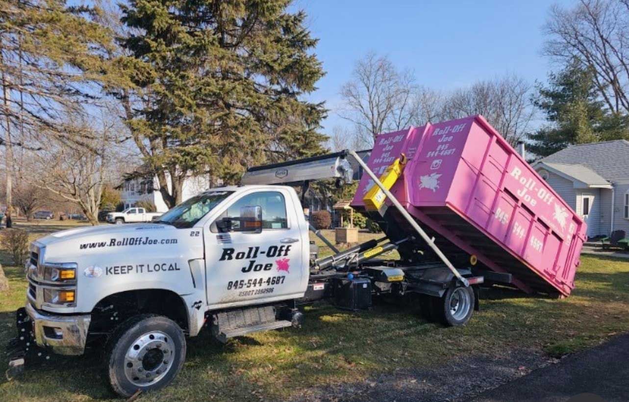 White truck with pink dumpster, parked on grass. The dumpster is tilted, ready to be unloaded, next to a house.