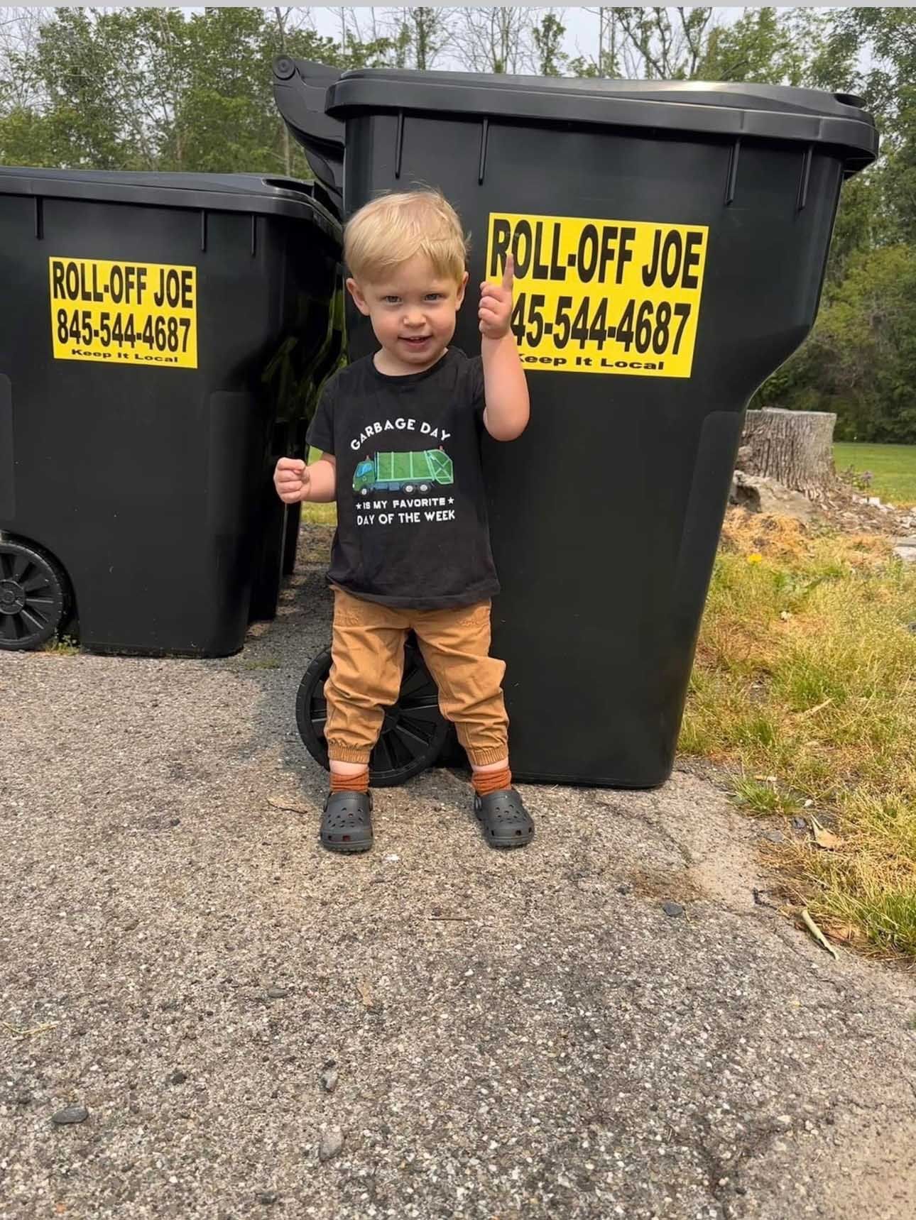 Young child stands between two black dumpsters, giving a thumbs up. 