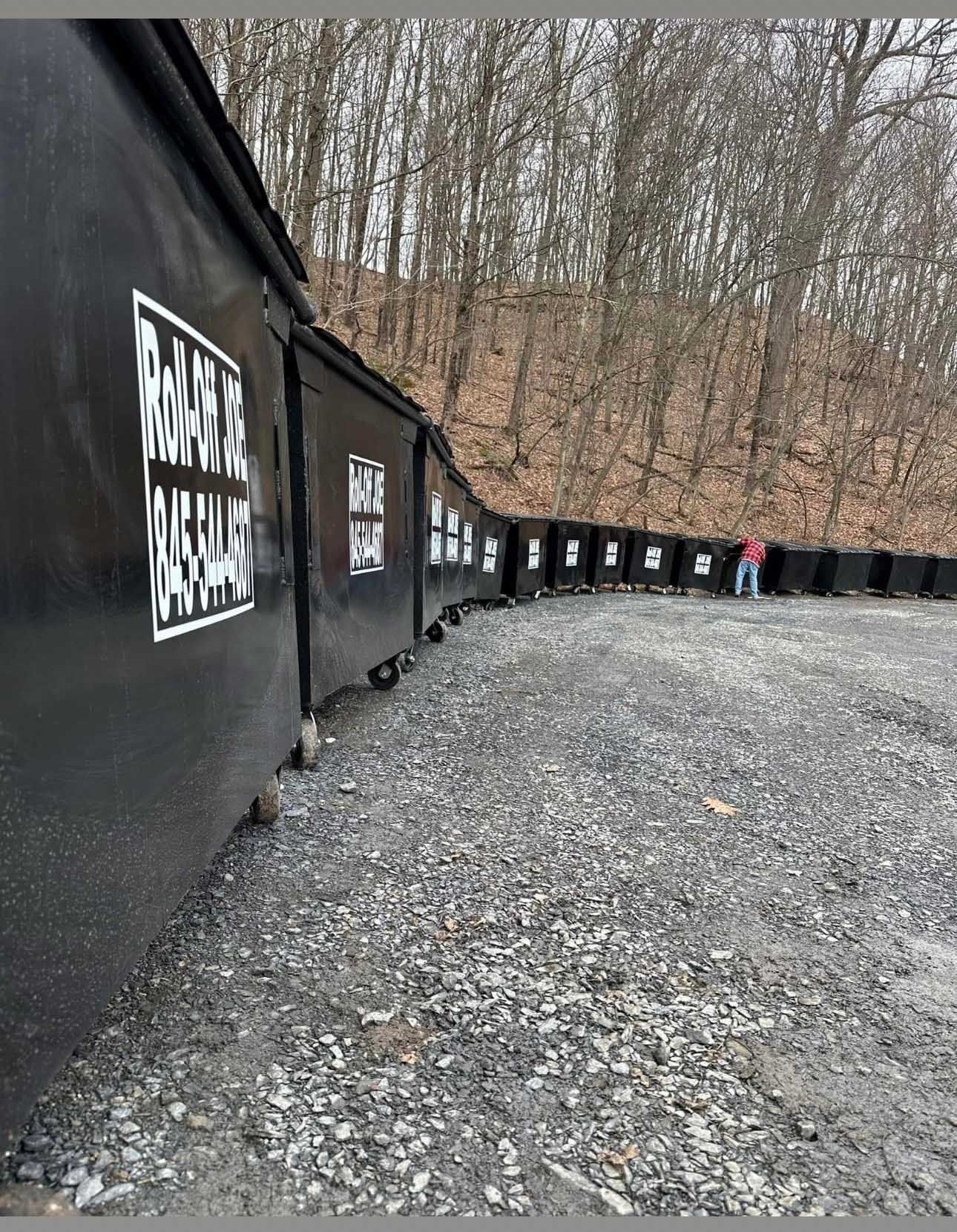 Dark train cars curve along a gravel path. A person stands near the end. Trees line the hillside.