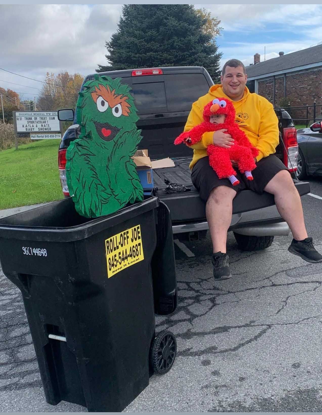 Man in yellow hoodie holding Elmo, with Oscar the Grouch and a trash bin in front of a truck.