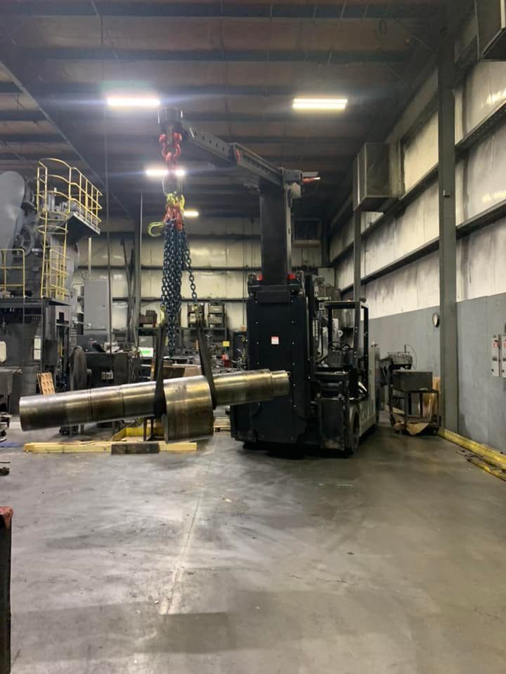 A large metal cylinder is lifted by a crane inside an industrial warehouse. A forklift supports the crane.