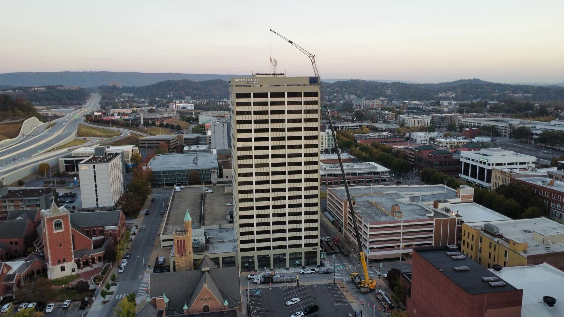 Aerial view of a tall building under construction in a city. A crane is on top, and other buildings and roads surround it.