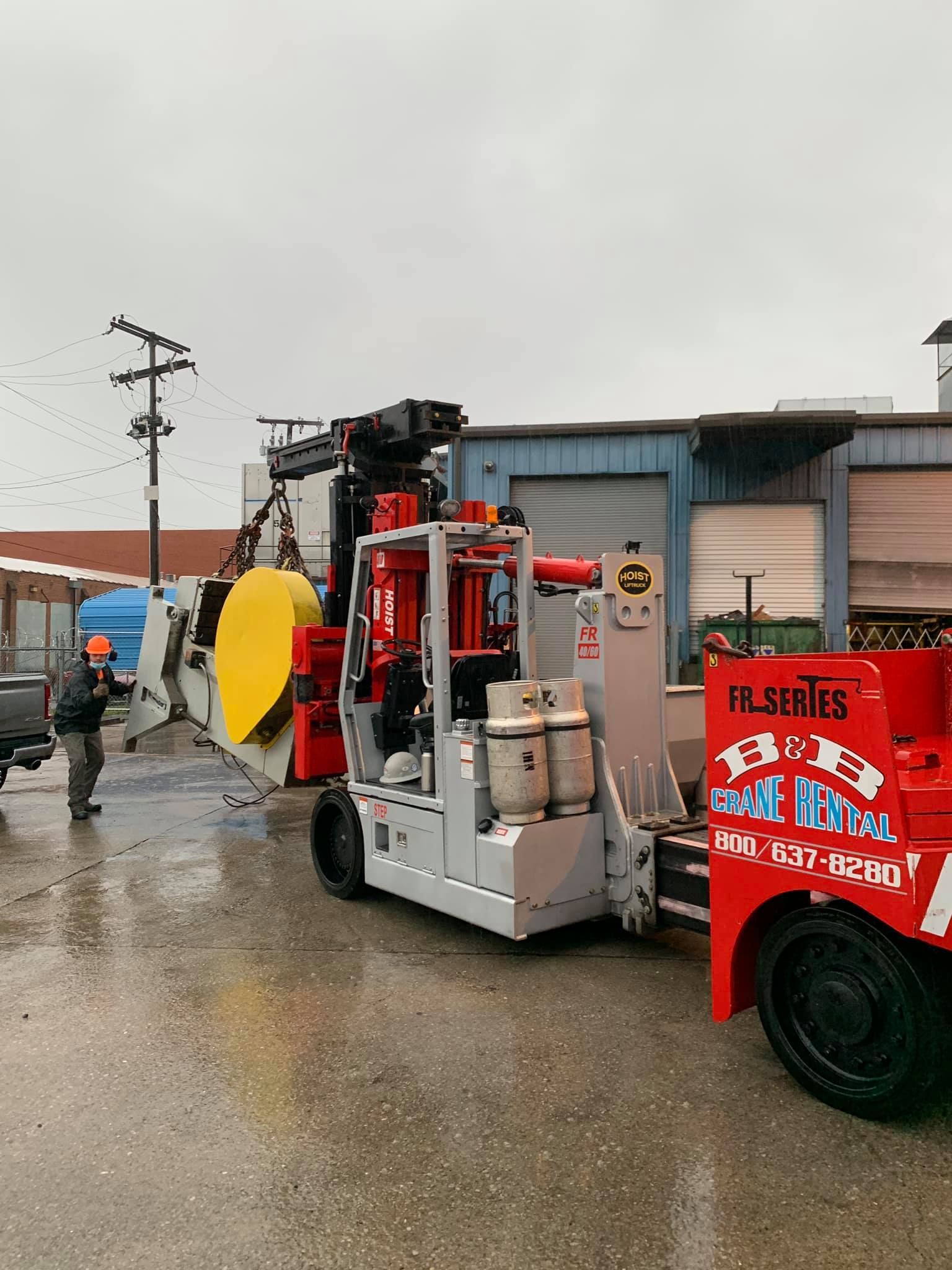 A red truck is towing a forklift in a parking lot.