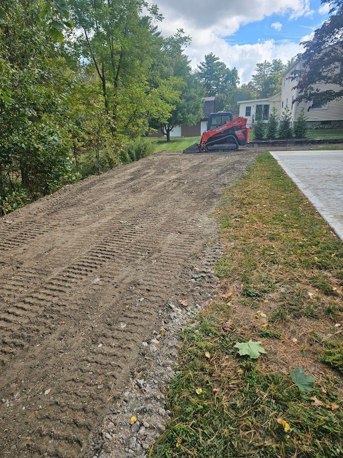 A red skid steer loader sits on a dirt surface being graded next to a paved driveway and trees under a cloudy blue sky