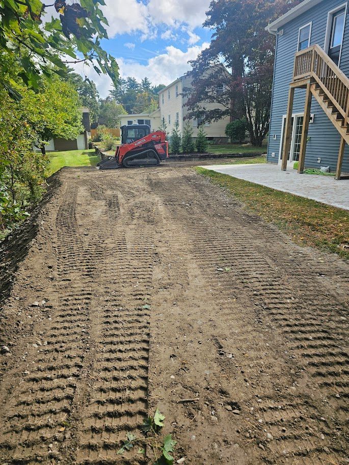 A red skid steer loader sits on a dirt plot next to a blue house with wooden stairs on a sunny day