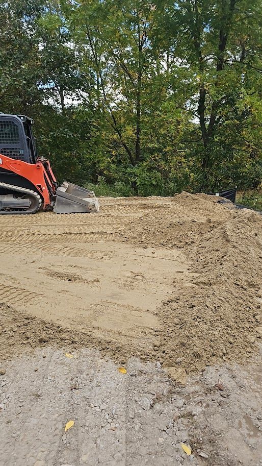 An orange skid steer loader parked on a dirt patch near a treeline, with tracks visible in the loose soil