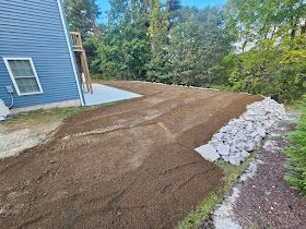 A freshly graded, dirt-covered yard next to a blue house, bordered by a line of large, grey landscape rocks