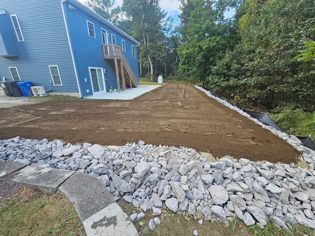A freshly graded dirt yard beside a blue house, bordered by a large stone retaining wall and landscape fabric