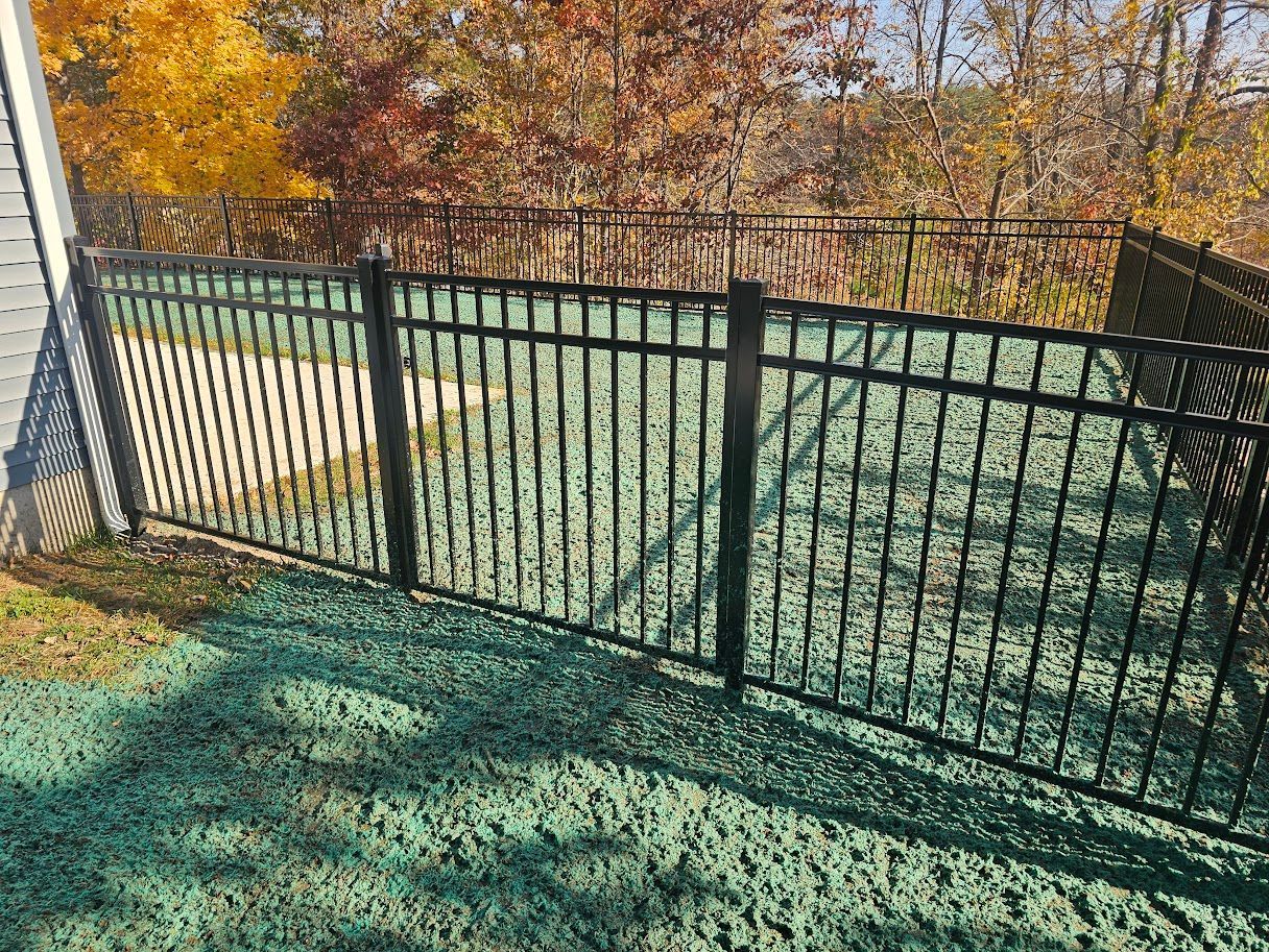 A black metal fence encloses a backyard area covered in green hydroseed mulch, with autumn trees in the background