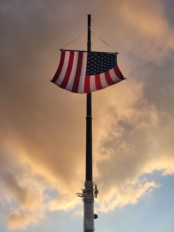 An American flag hangs horizontally from a tall pole against a cloudy, sunset-lit sky