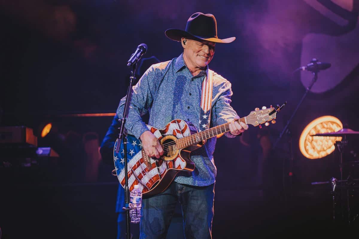 A musician in a cowboy hat playing a guitar painted with an American flag on a stage with purple lighting
