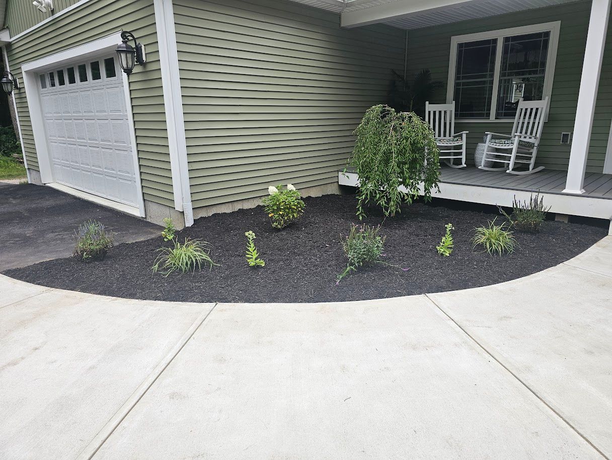 Landscaped garden bed with mulch and plants in front of a house with a garage and porch.