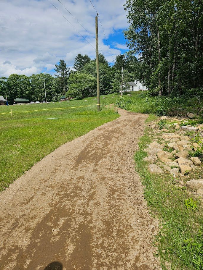 A wide dirt path curves through a grassy field toward a wooded area with a utility pole and building in the background.