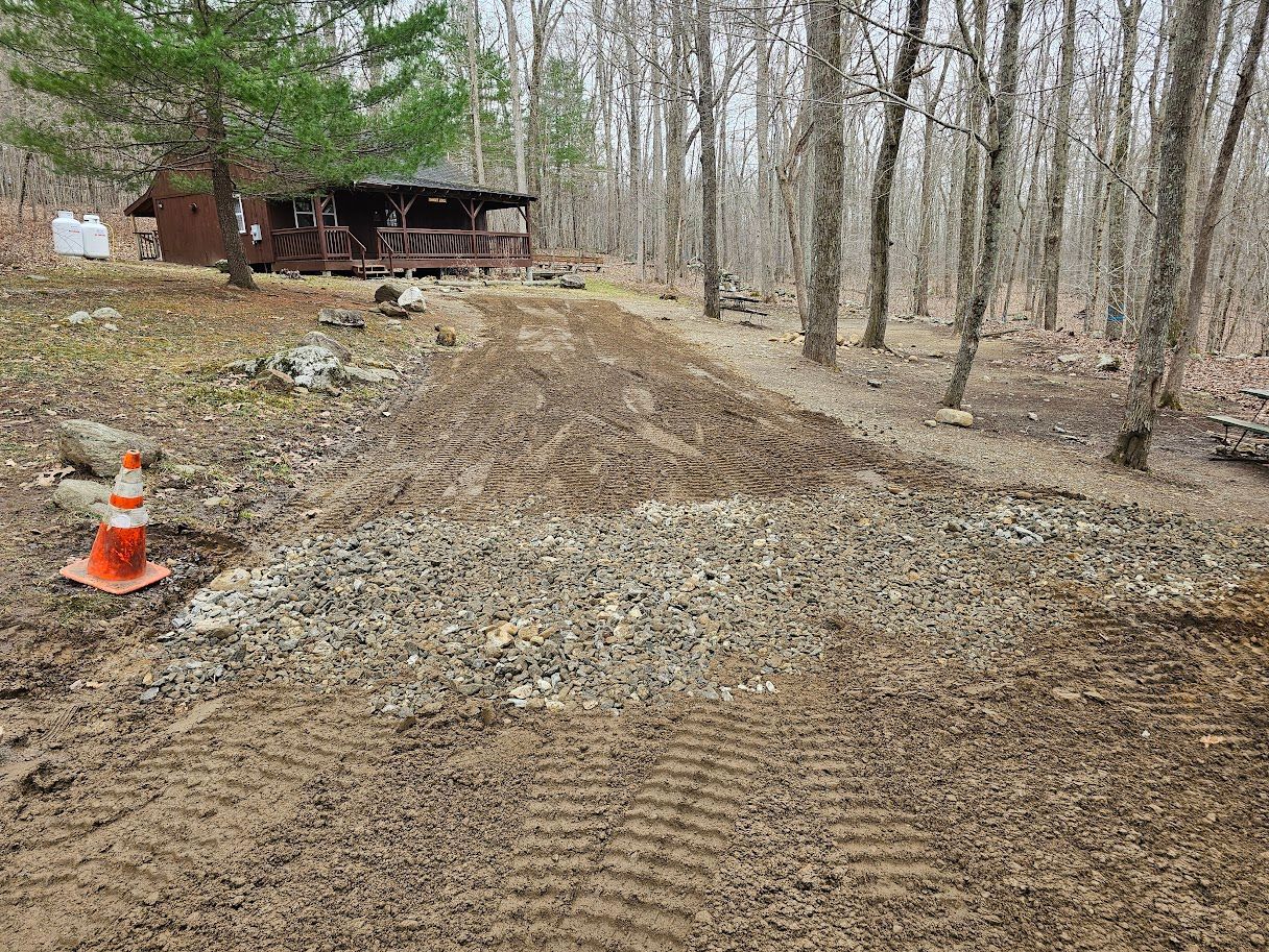 A cleared dirt path with patches of gravel leading toward a wooden cabin in a wooded area, with a traffic cone nearby.