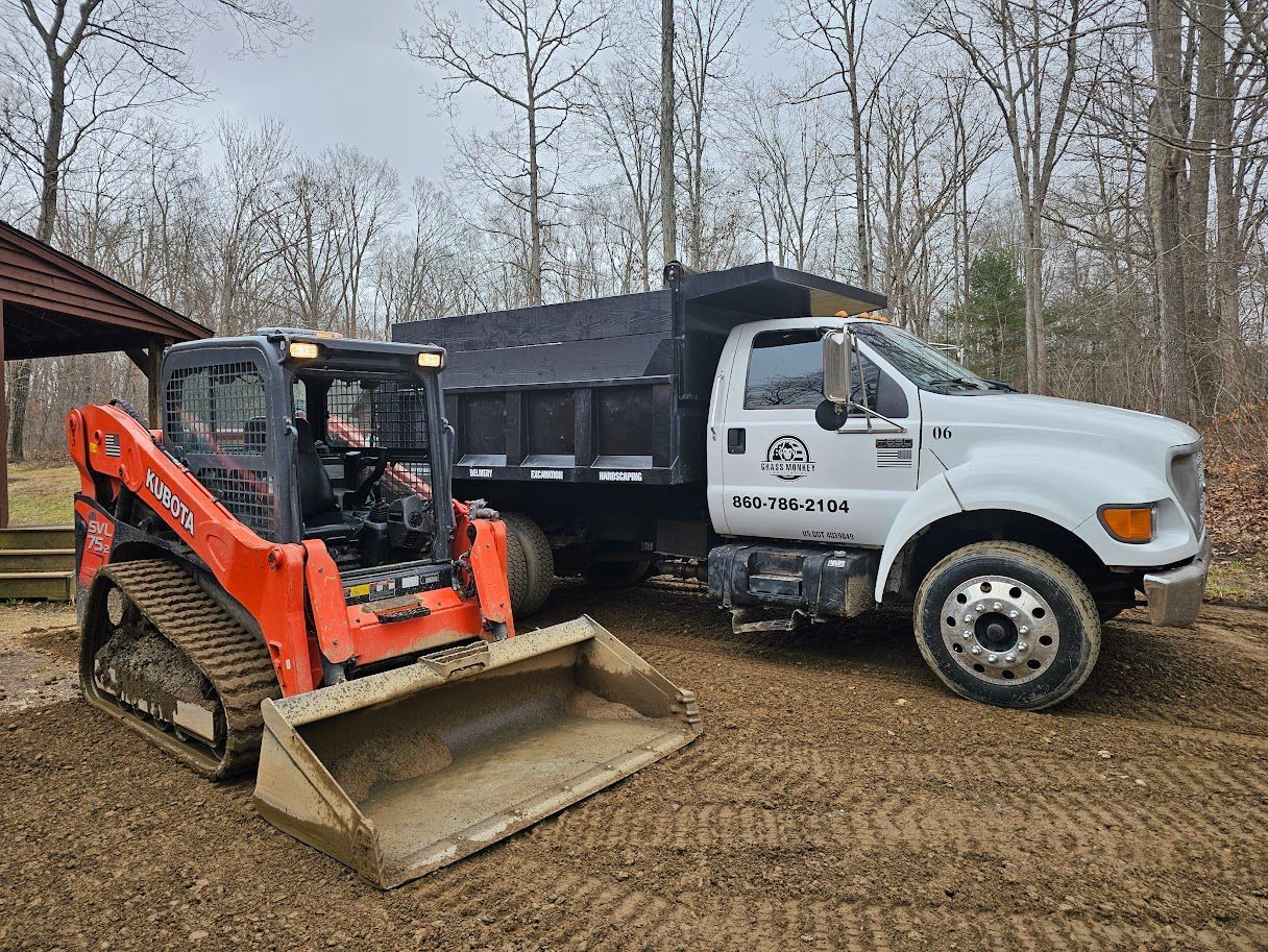 An orange track loader sits in front of a white dump truck on a dirt lot, with trees in the background.