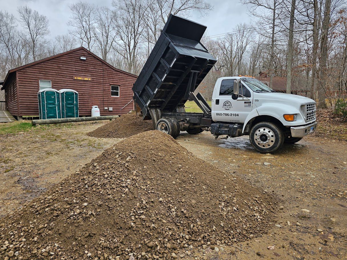 A white dump truck with its bed raised, emptying a pile of gravel onto a dirt surface next to a brown cabin.