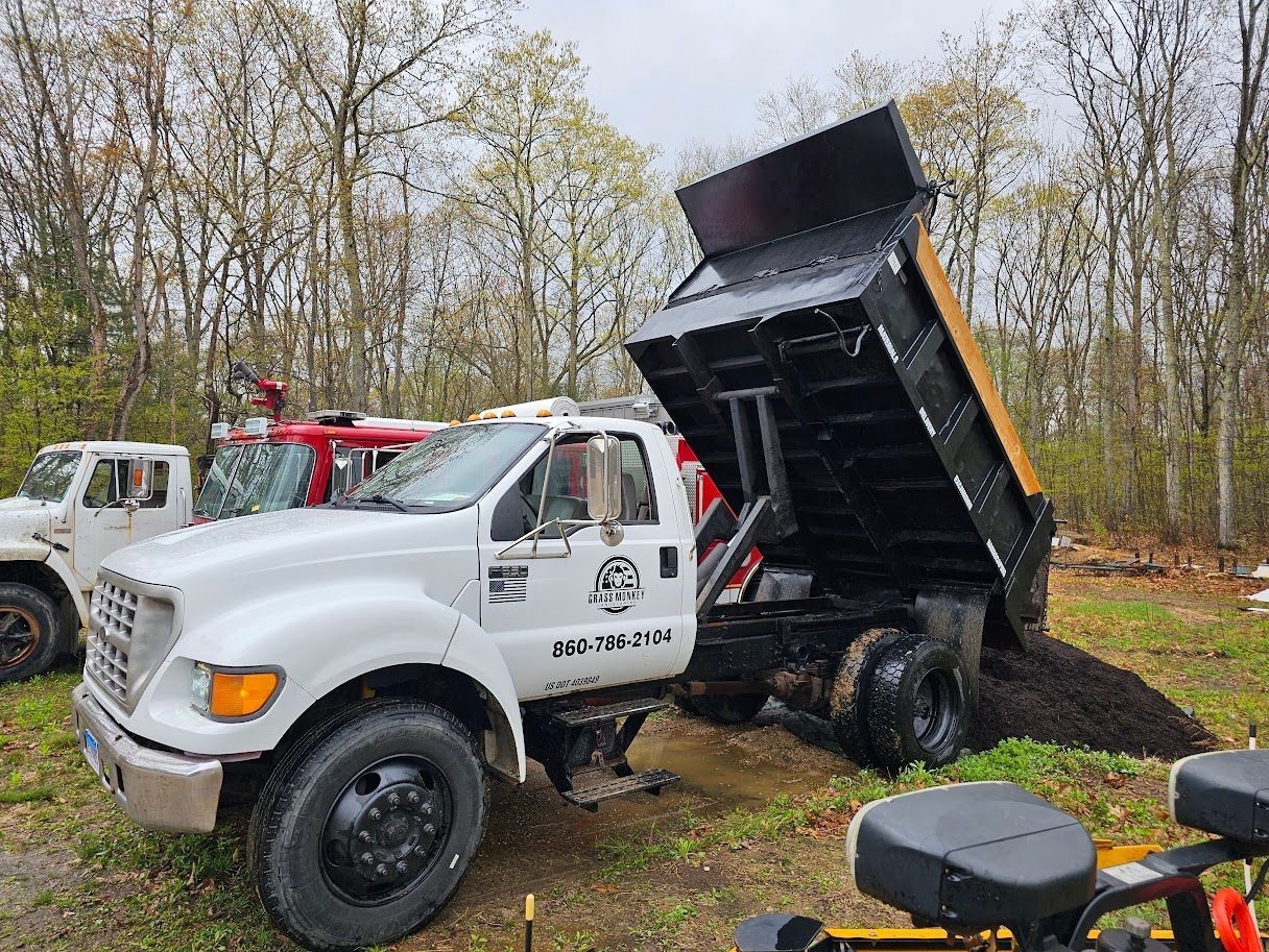 A white dump truck is parked in a wooded area with its black bed raised, dumping a pile of dark soil onto the ground.