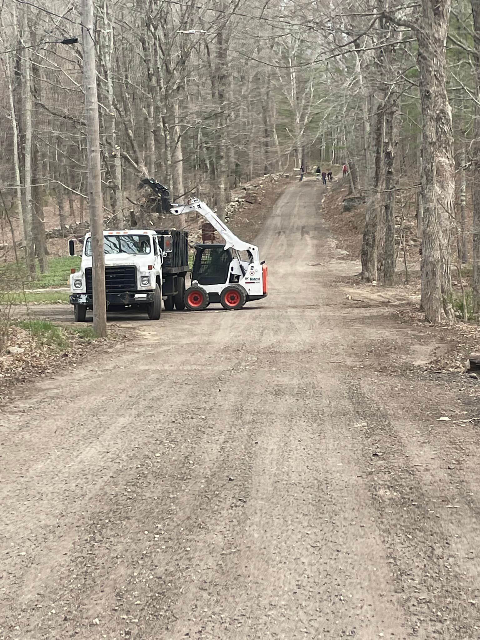 A white skid steer loader lifts a load into a white dump truck on a dirt road in a wooded area.