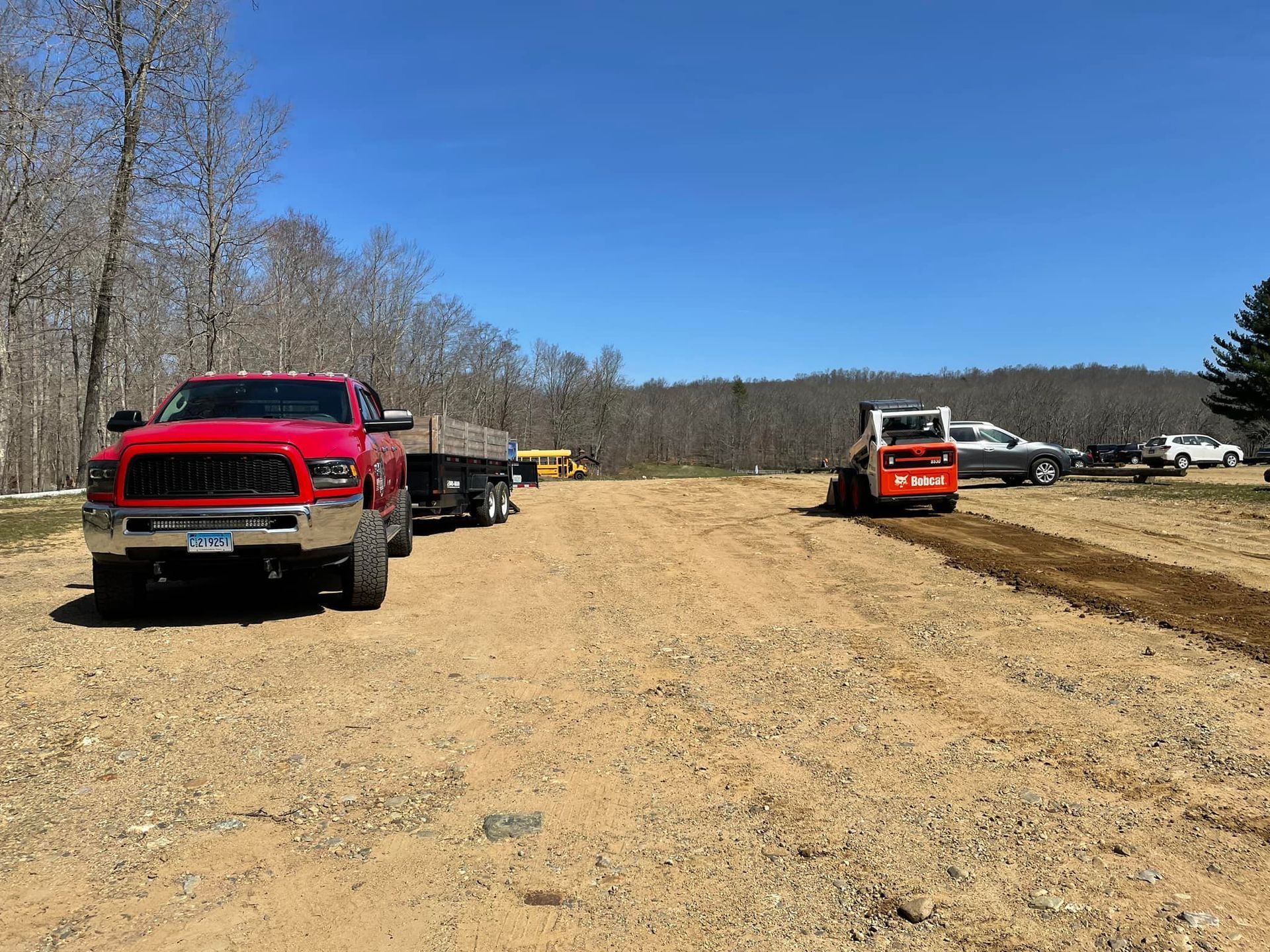 A bright red pickup truck with a trailer, alongside a Bobcat skid steer working on a gravel lot under a clear blue sky.