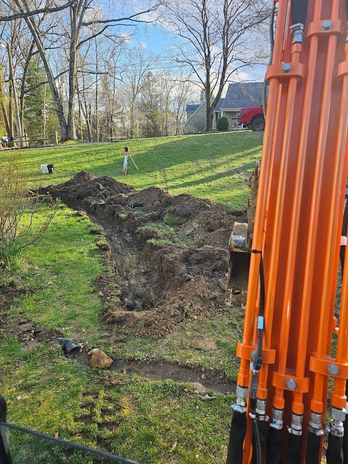 Excavator digging a trench in a grassy backyard, likely for utility lines. Orange machinery, trees, and houses in the background.