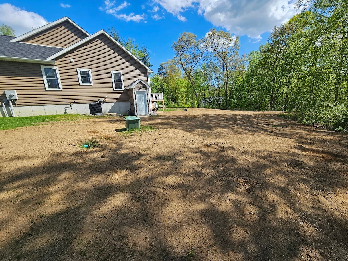 A cleared, brown dirt lot in front of a house, with green trees and a blue sky in the background.