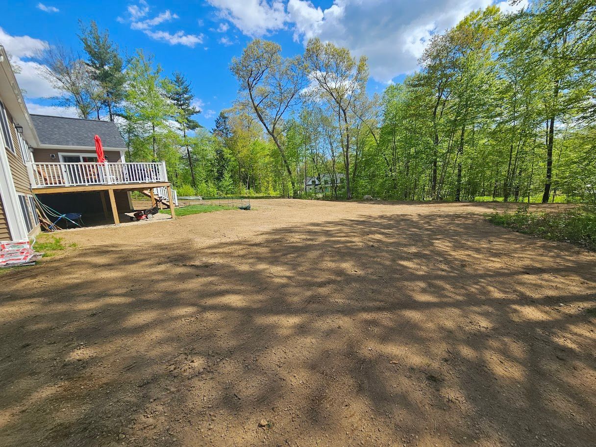 Cleared dirt area with a house and trees in the background under a blue sky.