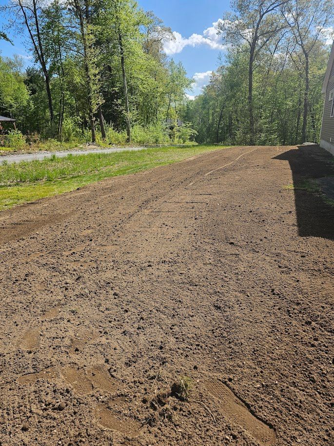 A newly tilled brown plot of land in front of a green grassy area and trees on a sunny day.