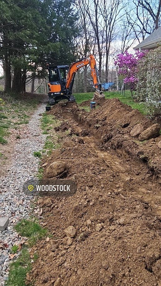 Orange excavator digging a trench in a yard, trees in the background, a house on the right.