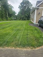 Green lawn in front of a tan house. A car is parked on the right. Trees in the background.