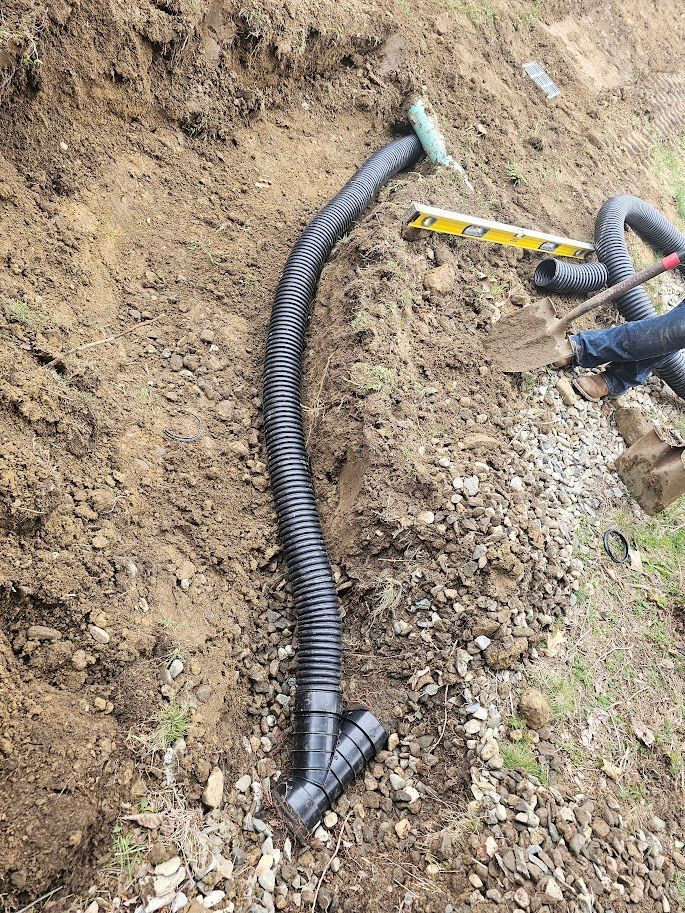 Black corrugated drain pipe installed in a trench; a person is working nearby with a shovel.
