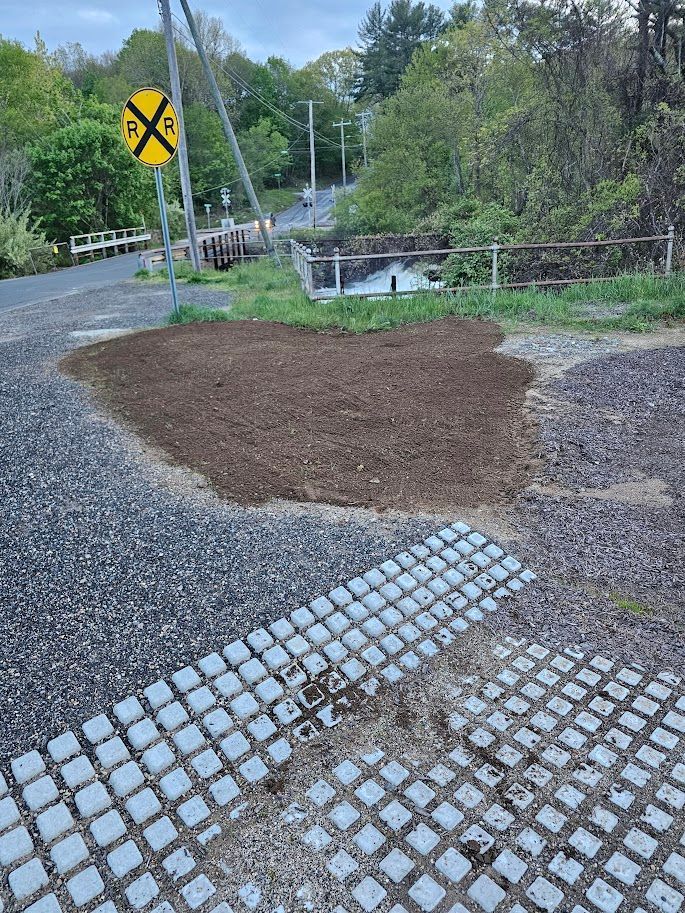 Gravel and dirt area near a railroad crossing with a warning sign, grassy vegetation, and a white bridge in the background.